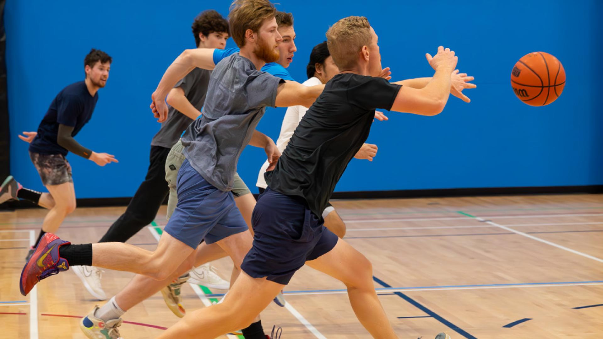 Basketball players chasing a ball in a Sportsplex gym.