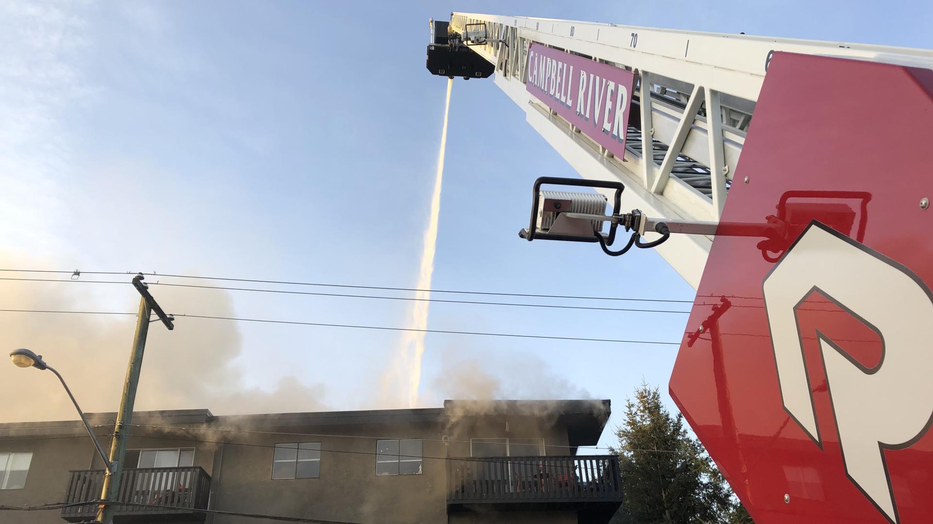 The ladder of a fire truck stretches into the sky, spraying water on a smoky apartment building.