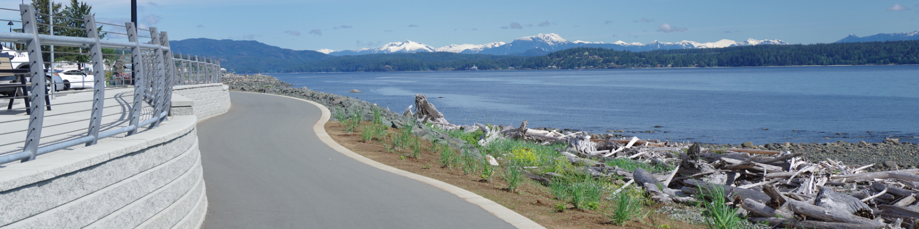 Cement walkway along the beach with driftwood in the foreground and the ocean and mountains in the background