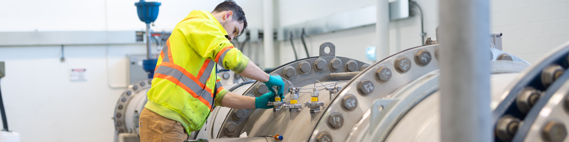 A person wearing safety gear working on large industrial pipes at a water treatment facility.