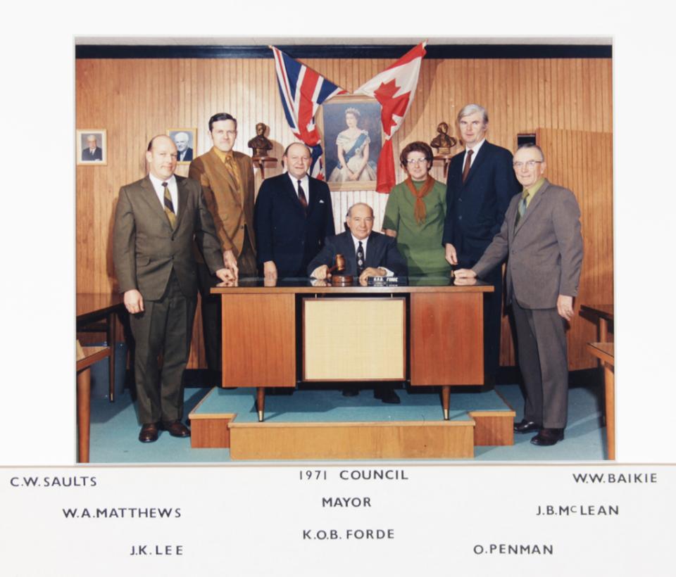 A group of eight people in formal attire around a table with flags in the background.