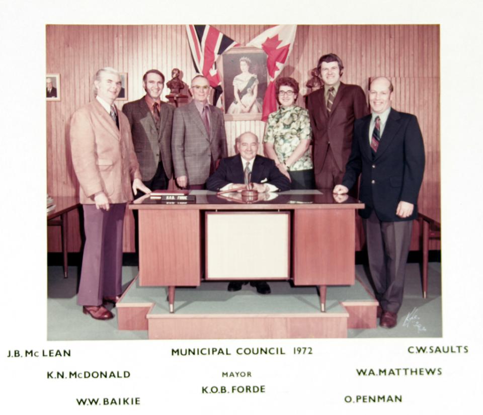 A group of men in formal attire pose around a desk with flags and a portrait backdrop.