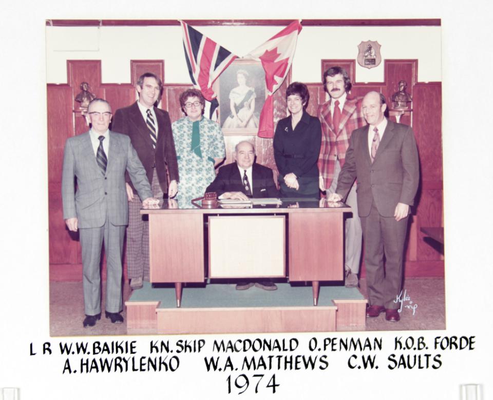 A group of seven people in formal attire stand behind a desk with flags in the background, 1974.
