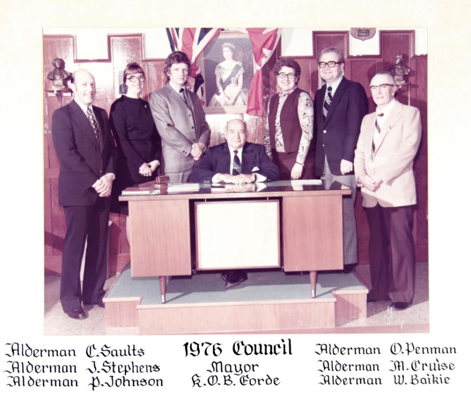 Council members pose around a desk, man seated in center, flags in background.