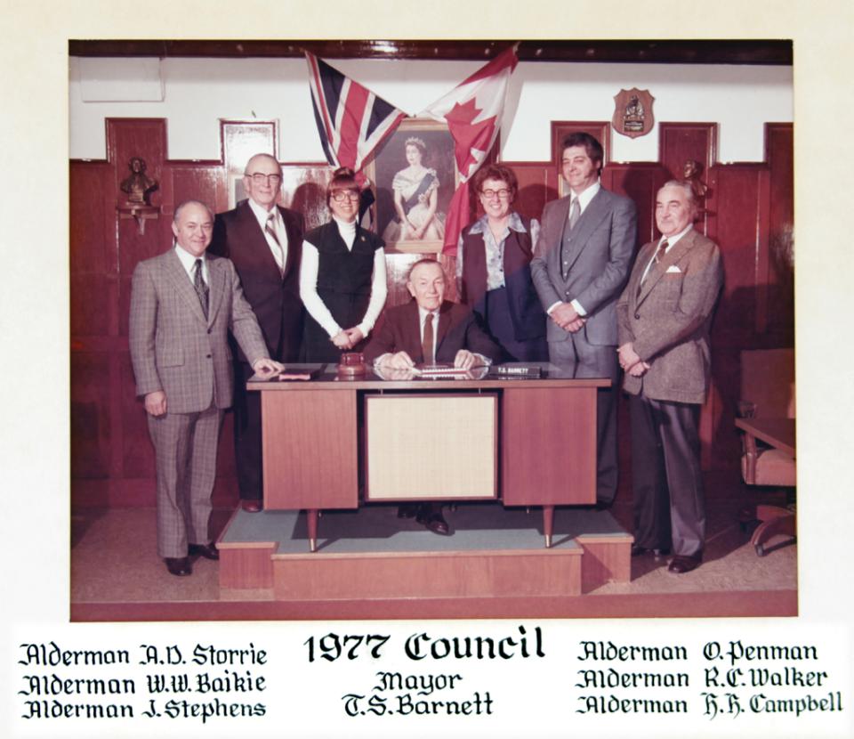 Group of nine people, one seated at a desk, in an office with flags and framed portraits.