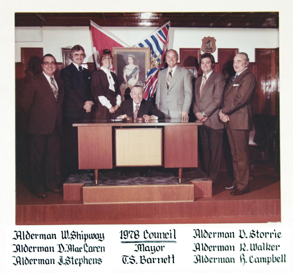 A group of officials in suits pose around a desk with flags in the background.