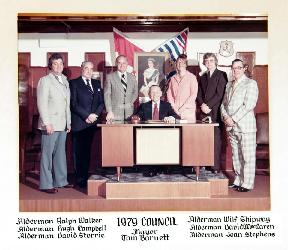 A group of men poses around a desk with flags in a council chamber.