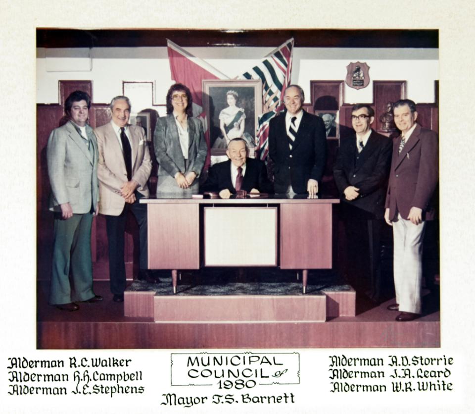 Council group portrait with people standing and sitting at a desk, flag in background.