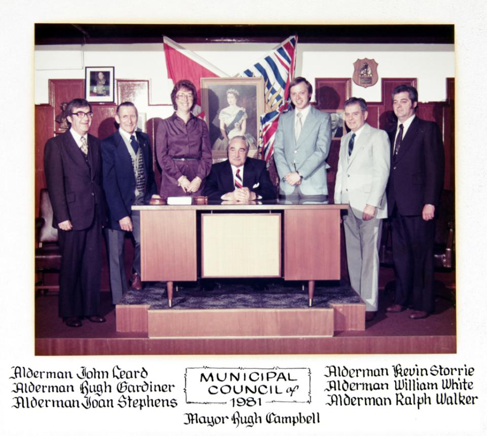 Seven officials in formal attire stand behind a seated mayor in a council chamber.