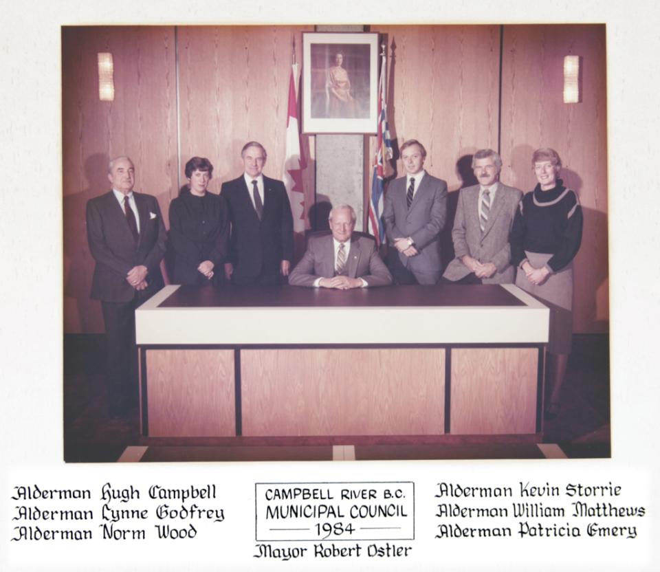 Mayor and council seated at a wooden desk, formal attire, large flags in the background.