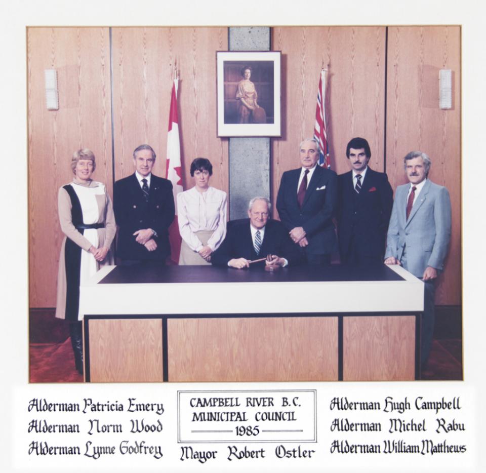 Council members posing with flags and framed portrait in office.