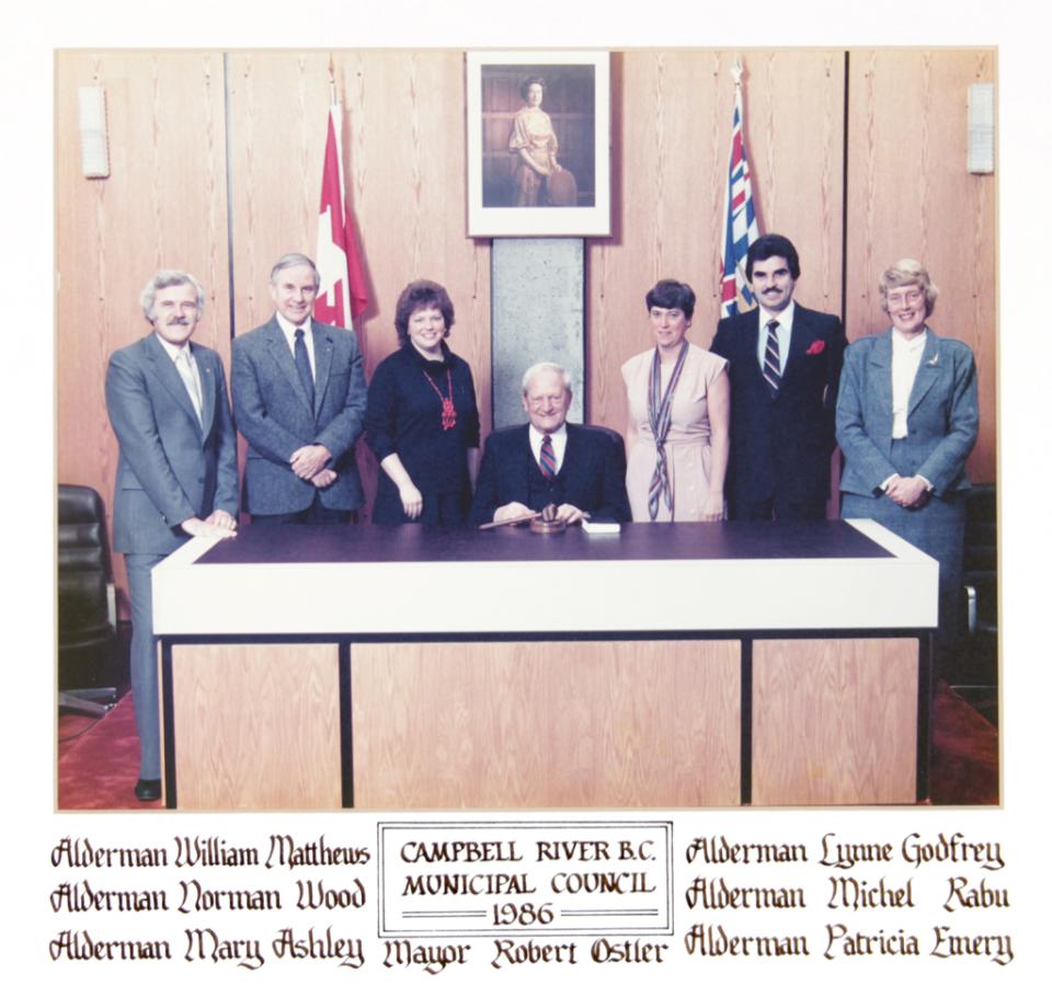 Council members standing behind a seated mayor at a desk, flags in the background.