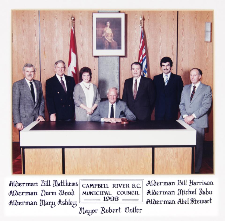 Seven people standing behind a seated man at a desk, in a formal setting with flags.