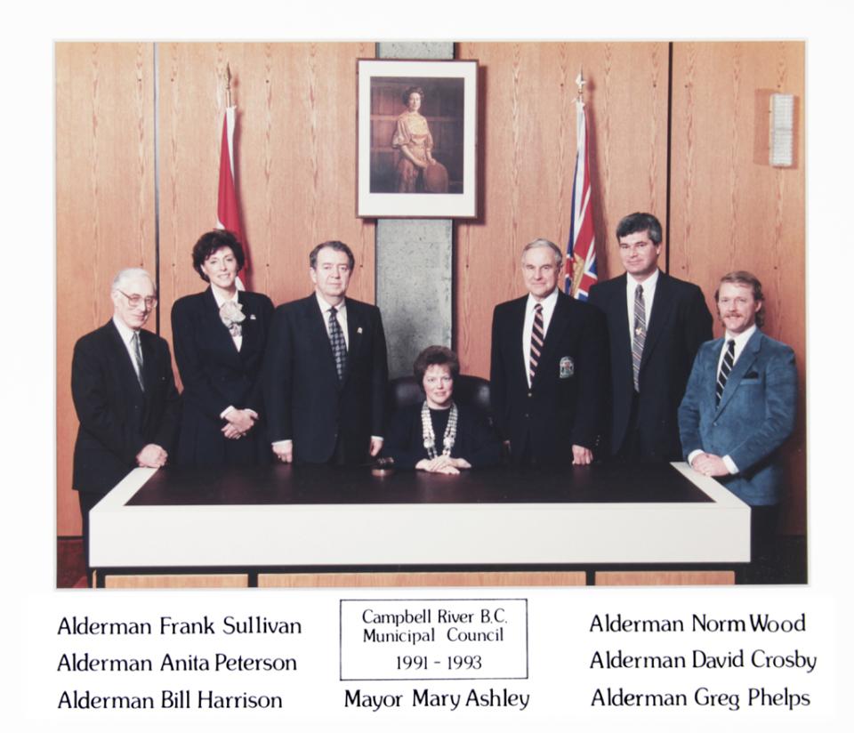 Council members pose in suits around a seated mayor in a wood-paneled room.