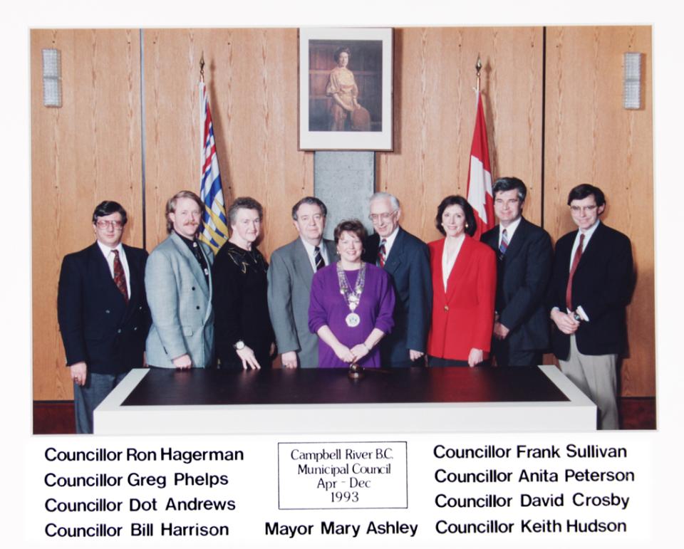 A group of nine people in professional attire stand in front of flags and a wooden wall.