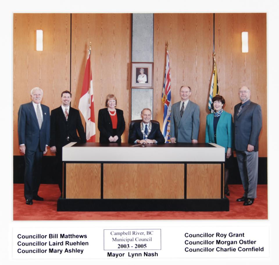A group of officials stands and sits around a table with flags in the background.