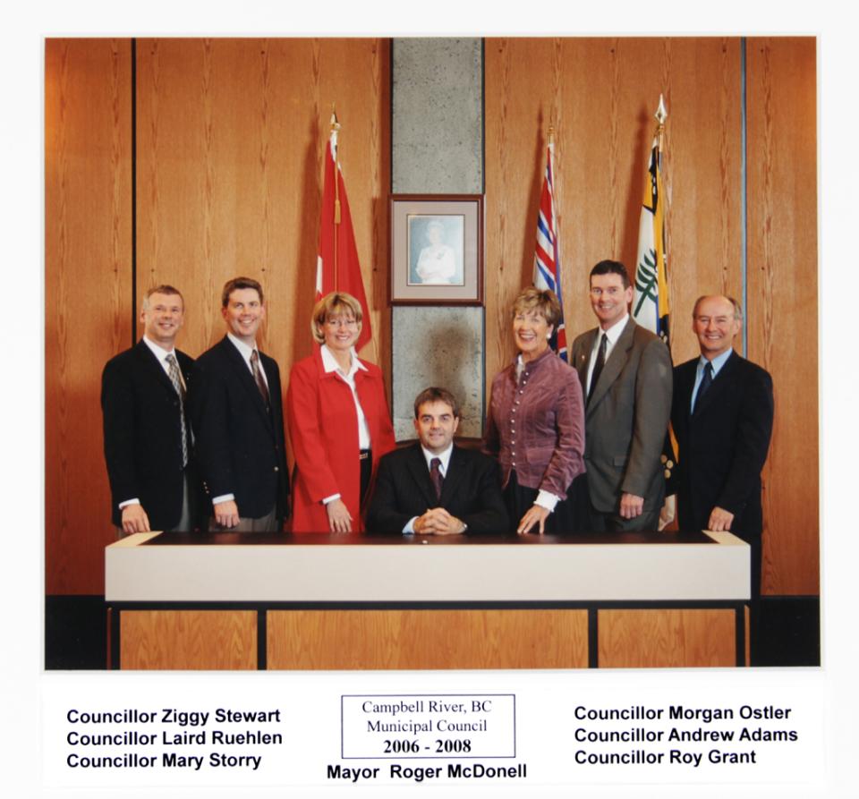 Seven people posing formally in a council chamber with flags behind them.