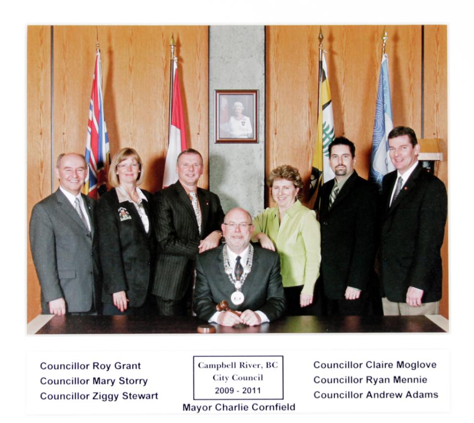 Group of council members standing behind a seated mayor in a formal setting.