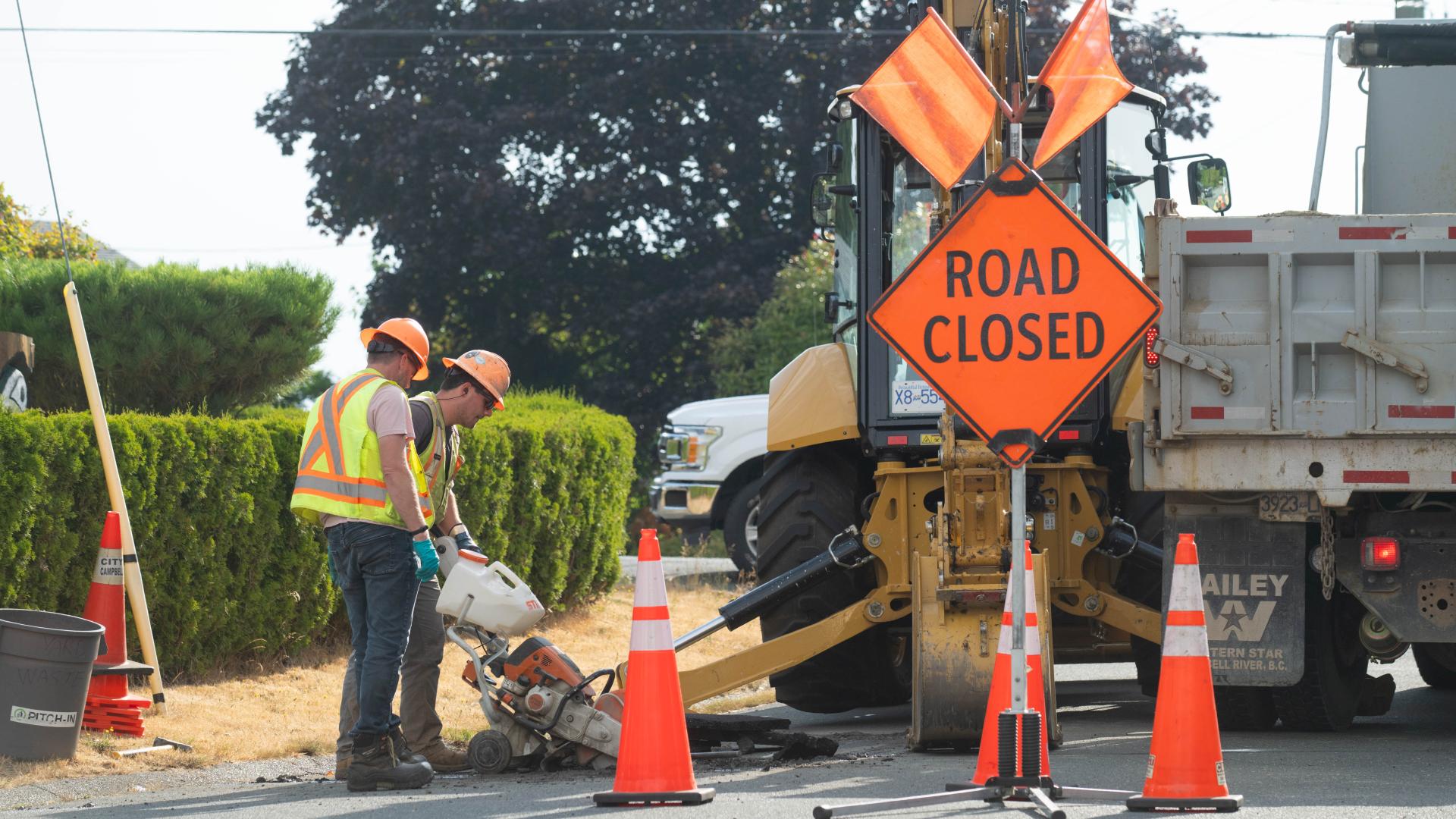 Workers operating machinery at a road construction site with a "Road Closed" sign.