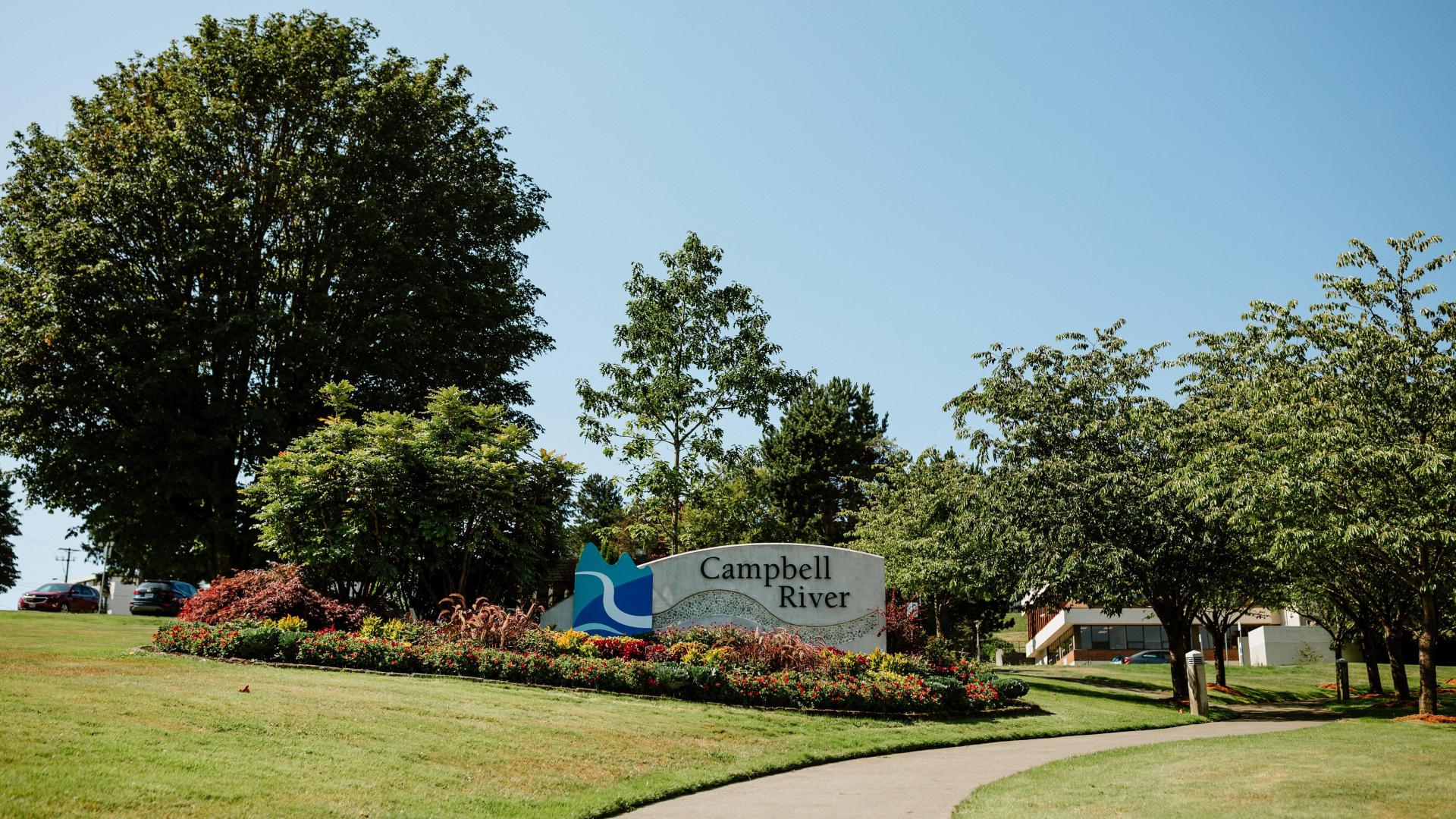 Sign at the front of the City of Campbell River City Hall surrounded by a flowers, trees and a path on the right side.