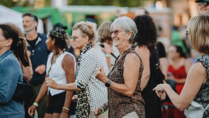 A crowd dances at an outdoor concert on a sunny day. They are smiling and laughing, and the atmosphere is festive.