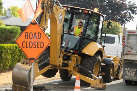 A person wearing safety gear, including a reflective vest, operates machinery in a construction zone. A bright orange sign reads "Road Closed".
