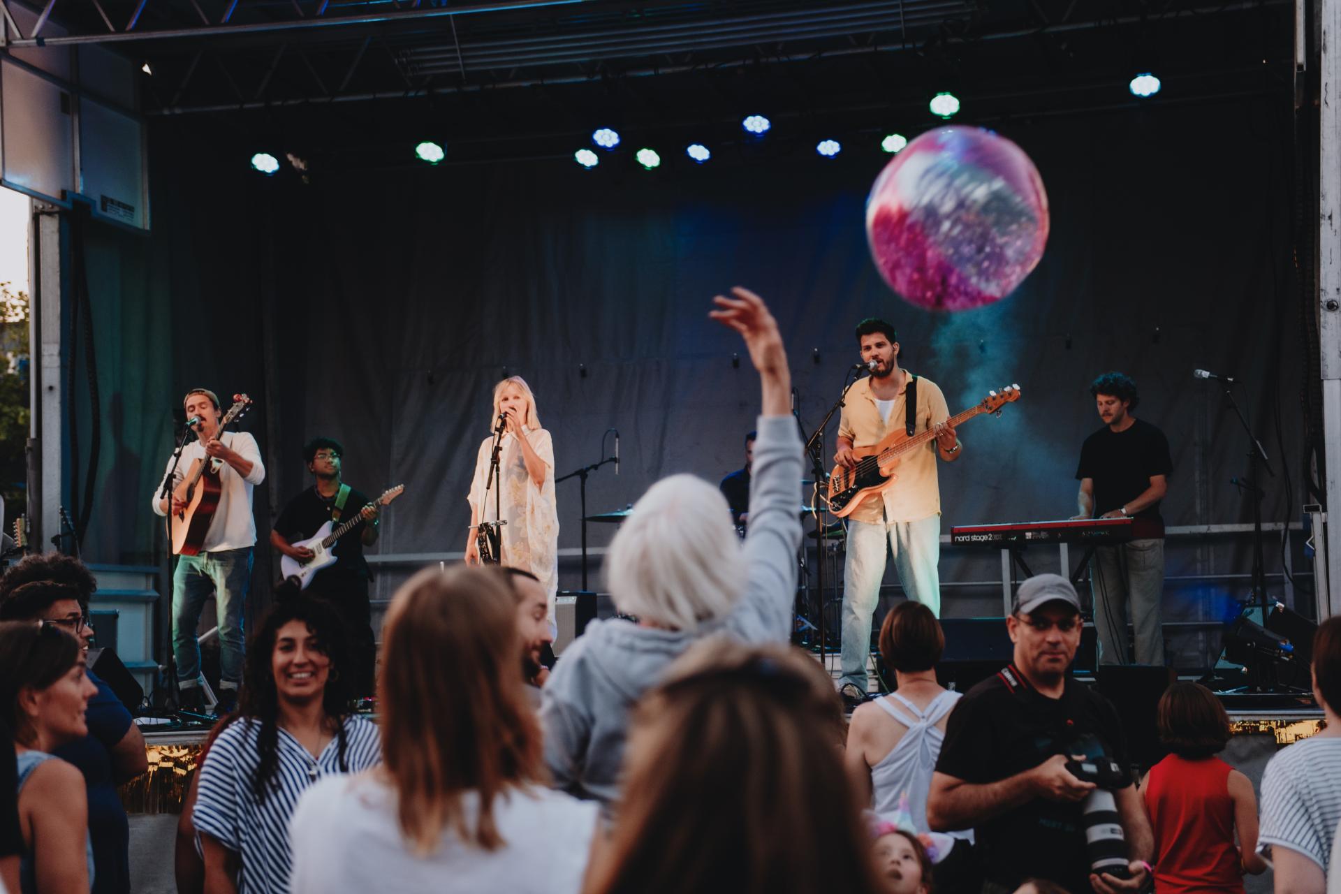 A band performs on an outdoor stage at CR Live Streets. An audience member tosses a colourful beach ball into the air.